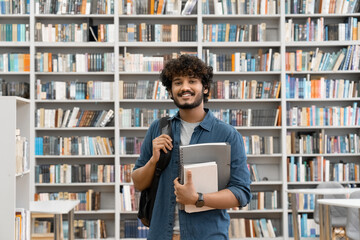 Young smiling Indian male student standing in university library with backpack