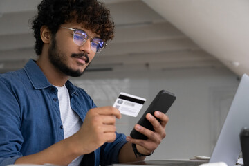 Young Indian man holding plastic bank card and smartphone make online payment