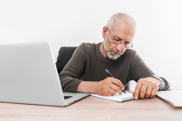Elderly man works at a laptop and writes in a notebook