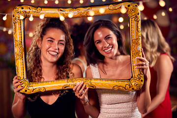 Female Friends Having Fun Posing With Photo Booth Photo Frame At Party In Bar - obrazy, fototapety, plakaty