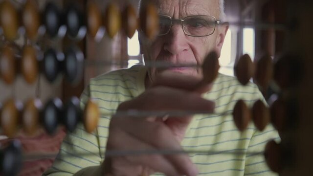 Mature accountant wearing glasses counts home finance with old wooden abacus on foreground. Calculation and estimation concept. Pension and retirement economy.