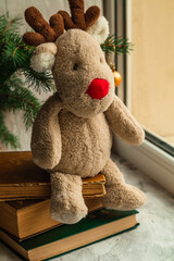 Christmas soft toy reindeer Rudolph sits on books near the Christmas tree and looks out the window.