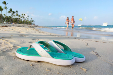Holidays on a tropical coast, beach flip-flops on sand on background of coconut palm trees, ocean and walking couple