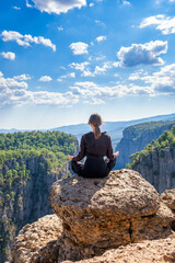 Naklejka na ściany i meble Back view of anonymous peaceful female sitting in lotus pose on edge of rocky cliff above mountains in highland during yoga session, vertical orientation, copy space