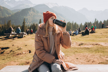 Young blond woman with thermos cup resting on the bench in mountains. Freedom, food, travel and vacations concept, outdoor activities. Happy Girl wearing red hat, halt