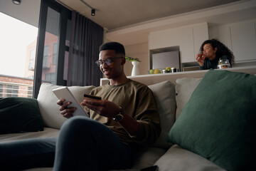 Young adult black male making online purchase on sofa with girlfriend in modern apartment