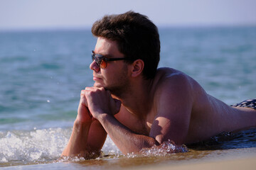 A young handsome man of 25-30 years old, a brunette, in weed sunglasses, lies relaxed on the sand on the beach of the sea coast and looks into the distance