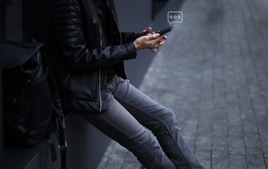 a young stylish man in a black jacket is texting via instant messengers on the phone near the office building.