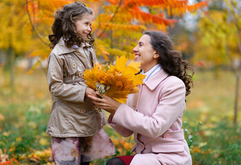 Mother and daughter posing in an autumn park. Beautiful yellow trees as background. They hug and are happy together.