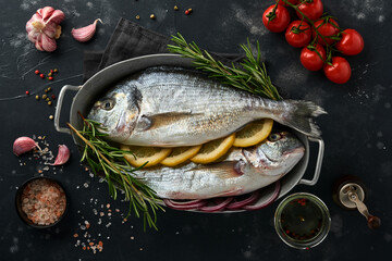 Raw dorado fresh fish or sea bream with ingredients for making lemon, thyme, garlic, cherry tomato and salt on a black slate, stone or concrete background. Top view with copy space.