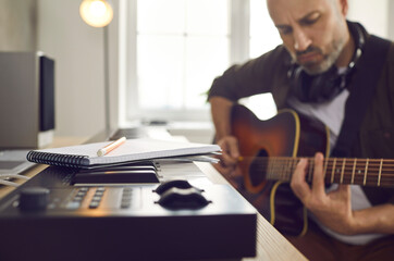 Musician and songwriter writing and playing music. Serious focused man picking guitar strings and thinking of new song while sitting with pencil and notebook by electronic keyboard in home studio - obrazy, fototapety, plakaty