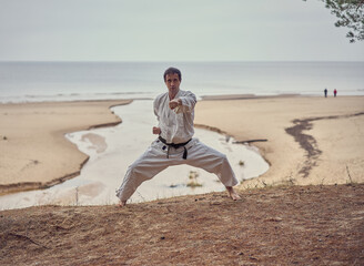 Fototapeta na wymiar Karate man in an old kimono and black belt training punch in kibadachi stand at the sea. Martial arts concept. The river flows into the sea at the background. 