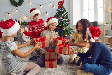 Group of happy children in Santa hats giving presents to their friend or sister on Christmas morning in cozy living-room. Happy girl receiving lots of New Year gifts from her generous friends