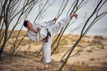 Karate man in an old kimono and black belt training side kick at the sea. Martial arts concept. The...