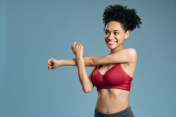 Portrait of confident young slim woman stretching her arms while standing isolated over blue background at studio and looking away