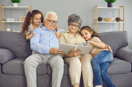 Happy grandparents and their two granddaughters look at a photo book with a family photo together. Grandparents and little girls at home sitting on the couch in the living room. Family concept.