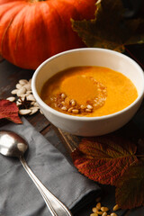 A bowl of homemade pumpkin soup with fresh pumpkin and gray kitchen cloth on white table with autumn leaves.