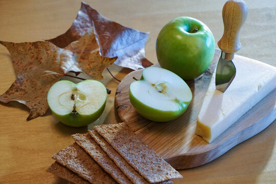 Autumn cheese board with crisp green apples and whole-grain crackers on wood table decorated with seasonal leaves.