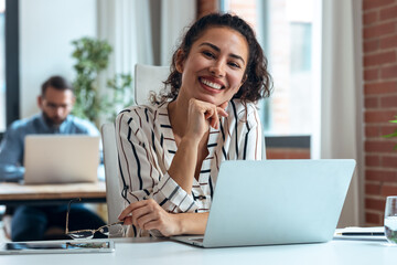 Smiling business woman working with laptop while looking at camera in modern startup office.