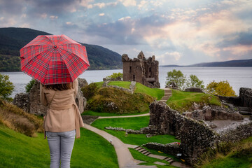 A tourist woman with a scottisch pattern umbrella looks at the famous Urquhart Castle at Loch Ness...