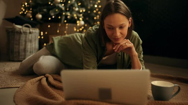 Happy young woman with cup of tea using laptop while laying near the Christmas tree. Attractive young woman texting on laptop. High quality 4k footage