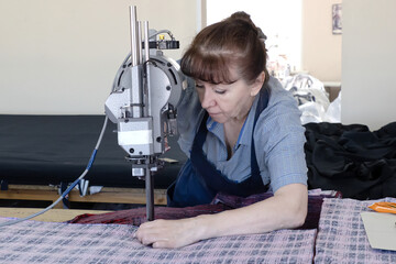 Female worker on a sewing manufacture uses electric cutting fabric machine. Textile industry,...