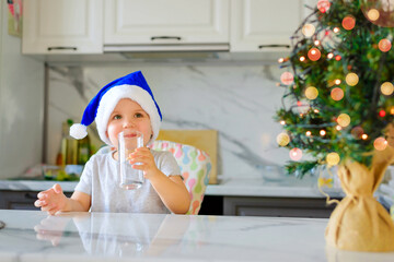 Cute boy kid in a blue Santa hat drinking filtered water from a glass in the kitchen. Holidays, health concept.