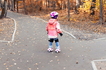 child girl in a pink helmet rides in the autumn park on four-wheeled rollers