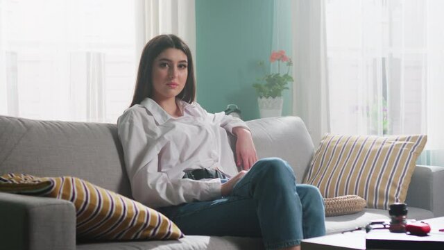 Portrait of young confident woman, dark-eyed brunette in white shirt, sitting on sofa and looking on camera, successful and self-assured, Slow motion.