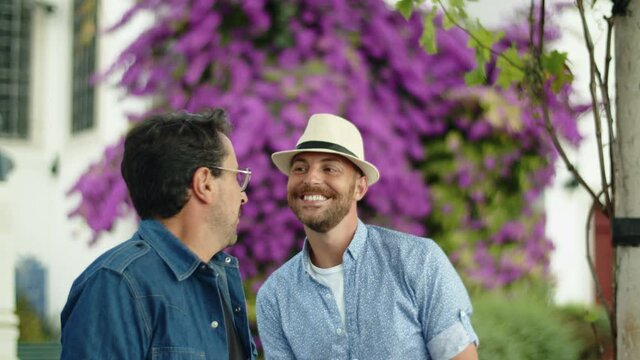Slow motion of gay in hat flirting with his smiling boyfriend. Medium shot of handsome Caucasian homosexual men having date in public garden, talking, spending time together. LGBT, love concept