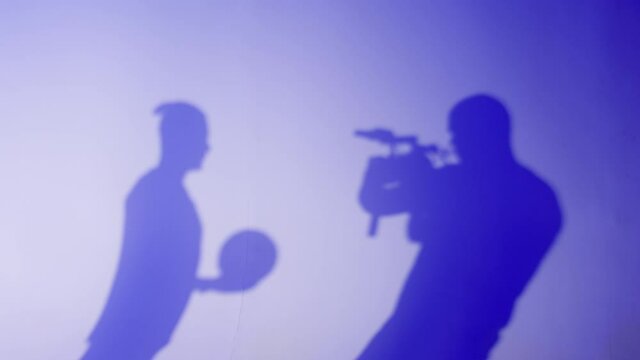 Shadows of football player holding ball, backstage. Shooting sportsman portrait, training soccer in studio. Sports school concept. Young man playing, practicing on blue neon background. 