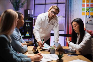 Caucasian man in white shirt looking on camera during conference with multiracial partners. Business people sitting at desk office with papers and modern technology.