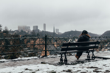 Woman sitting on a bench looking at a snowy city on a cloudy day with buildings in the background