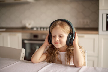 Naklejka premium Smiling cute little girl at home in headphones listens to music, audio. Child in the kitchen . kids and gadgets, blogger, streaming. technology concept. Music lessons, classical music