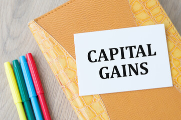 Text capital gains on a white card that lies on a leather notebook on the table next to colored markers. Financial concept