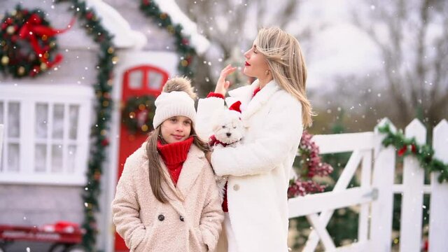 Beautiful blond woman in white winter coat holding small dog and posing with pretty young girl against Christmas ranch under the snowing. Slow motion.