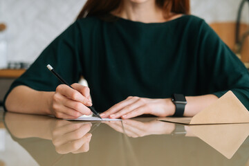 Close-up cropped shot of unrecognizable young woman writing handwritten letter sitting at table at home, selective focus, blurred background. Happy female writing wishes on Christmas card.