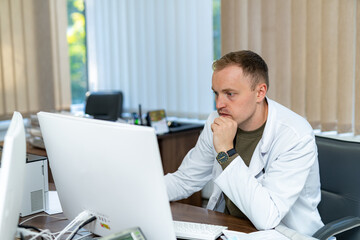 Handsome doctor in uniform working with computer. Attractive young medical specialist sitting with laptop.
