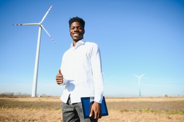 African engineer wearing white hard hat standing with digital tablet against wind turbine