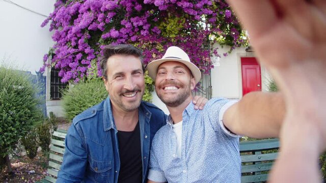 Handheld shot of homosexual men doing selfie in summer park. Front view of handsome Caucasian guys posing for camera, hugging, taking picture on phone during vacation in Europe. LGBT, date concept