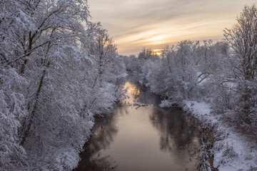 Small river running through the white snowy forest on frosty winter evening. Orange sunset sky in the background