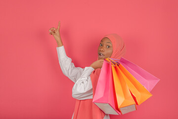 Profile of an excited young Muslim woman pointing fingers and holding many bright shopping bags looking at the camera on a pink background.