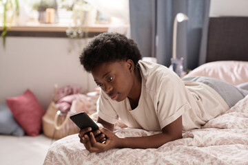 Fototapeta premium African young woman lying on the bed and watching something on her mobile phone in the room