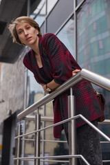 young woman with short hair in red checkered jacket outdoors in front of office building. portrait of caucasian female leaning on railings at street