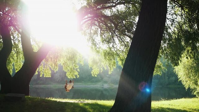 A child is swinging on a swing on a beautiful summer evening. The boy is swinging on a swing under a lush tree against an incredibly beautiful background of river. Sun rays break through tree branches