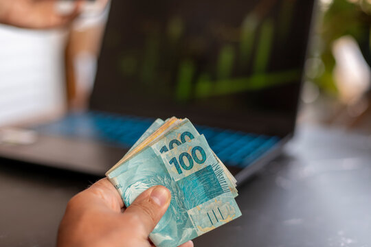 Brazilian money banknotes in hand, with blurred background of laptop computer
