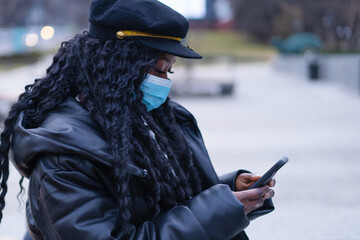 Young Afro girl with face medical mask uses the phone in a outdoors. African girl using facemask...