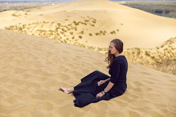 Fototapeta na wymiar woman in black long dress sit through the desert dunes in summer with her back