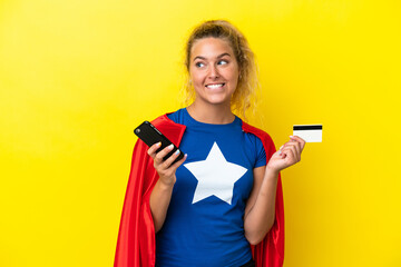 Super Hero woman isolated on yellow background buying with the mobile with a credit card while thinking