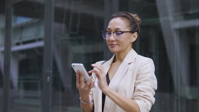 Attractive and successful hispanic business woman in elegant suit and fashionable eyeglasses scrolling web pages on smartphone, checking her schedule, sending important letters or receiving emails